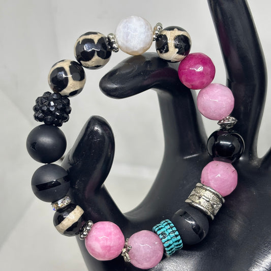 A beaded stretch bracelet displayed on a black hand-shaped jewelry stand. The bracelet features matte black beads, pink beads, leopard-print beads, a white marble bead, a textured black bead, silver spacer beads, and a turquoise-and-black woven accent bead.